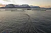 Laksemærer og fôrlinjer i sjøen ved Flakstadvåg på Senja med snødekte fjell bak.