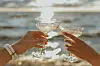 To hender skåler med champagneglass foran glitrende vann.