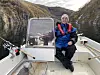 Person sitter i liten båt med fiskestenger i et smalt fjordområde omgitt av bratte fjell.