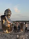 Tre arbeidere ved en stor metallskulptur av en menneskelig overkropp i et steinete landskap.
