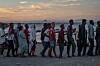 En gruppe migranter står i kø på en strand ved sjøen ved solnedgang.