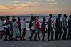 En gruppe migranter står i kø på en strand ved sjøen ved solnedgang.