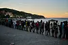 Lang kø av mennesker på en strand i Ceuta ved solnedgang, med havet og en ås i bakgrunnen.