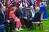 Kong Harald og dronning Sonja sitter i samtale med flere personer på en gressplen, med norske flagg i bakgrunnen.