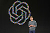 Sam Altman står på en mørk scene foran OpenAI-logoen.