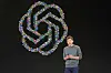 Sam Altman står på en mørk scene foran OpenAI-logoen.