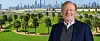 Smilende mann i mørk jakke foran en grønn park og en moderne skyline.