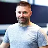 Smilende mann i grå T-skjorte foran en mørk bakgrunn.